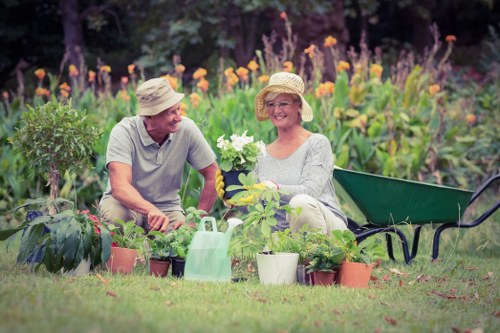 A couple of seniors enjoying gardening together in a well-maintained backyard garden, with lush green grass and a variety of potted plants and flowers arranged nearby. The man is wearing a light-colored T-shirt and a straw hat, while the woman is dressed in a light gray sweater and a straw hat, both smiling warmly. The woman is holding a white flowering plant, and in front of them are multiple small terracotta and plastic pots containing different plants, with a green watering can placed among them. To the right, there is a green wheelbarrow positioned on the grass, indicating ongoing gardening activities. Behind them, a dense, colorful flowerbed with tall purple, orange, and yellow flowers, possibly lavender or similar varieties, creates a vibrant backdrop, suggesting a landscaped outdoor space designed for leisure and horticulture. The scene is set in natural daytime light under clear weather, emphasizing the healthy foliage and floral elements, reflecting professional gardening and outdoor maintenance work associated with services like lawnmowing and garden care in the Shortlands area, near Bromley in south-east London.