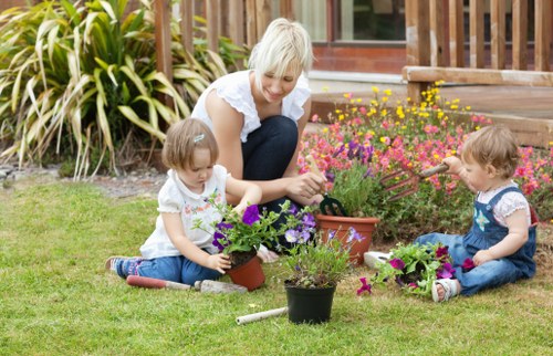 A woman with short blonde hair engaged in gardening activities in a well-maintained backyard, accompanied by two young children. The garden features a lush, green lawn with evenly cut grass, bordered by a variety of flowering plants and shrubs, including pink and purple blossoms. The children are seated on the grass, each holding small pots with blooming plants, while the woman is tending to one of the pots with gentle care. In the background, there is a wooden decking area leading to a modern residential building with large windows, framed by decorative plants and paving stones. The scene is illuminated by natural daylight, indicating a bright and mild weather condition. The outdoor space appears tidy, featuring a mix of natural and landscaped elements, consistent with professional gardening and lawn care services typical of the Shortlands area. This image demonstrates outdoor family gardening, with elements such as flower beds, lawn, and garden pots aligning with the services offered by Lawn Mowing Shortlands.