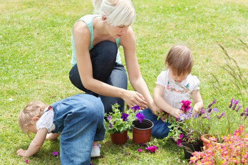 A woman with short blonde hair, wearing a light blue sleeveless top and dark jeans, is kneeling on a well-maintained, lush green lawn in a garden. She is engaged in planting or tending to a variety of potted flowers and plants, with several small terracotta and black plastic pots arranged around her. To her left, a young girl with light brown hair, dressed in a white top and blue jeans, is sitting cross-legged on the grass, carefully examining or handling flowers. Behind her, another small child with blonde hair, dressed in a white and patterned outfit, is leaning forward and closely observing the plants. The garden features a vibrant mix of flowering plants with purple, pink, and orange blossoms, bordered by a well-kept hedge and a paved pathway or patio edge in the background. The scene suggests a peaceful, outdoor space suitable for gardening activities, with natural daylight and a calm, sunny atmosphere that highlights the natural tones of the plants and the greenery. The image aligns with gardening and outdoor maintenance services offered by Lawn Mowing Shortlands in the South London area, enhancing garden aesthetics and outdoor enjoyment.